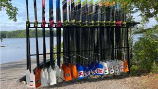 Oars with college insignia are linked up on a rack with Lake Wheeler in the background.