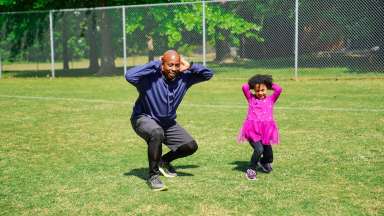 an image of a father and his daughter squatting in a field with their hands on their head