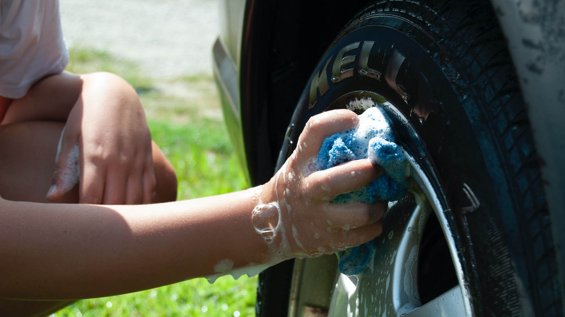 A person using a soapy cloth to wash a tire rim on the grass. 