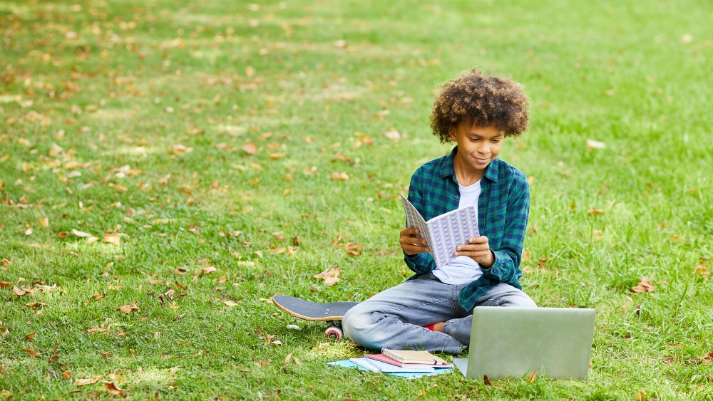 Teenage boy sitting out in grass holding notebook and looking at laptop