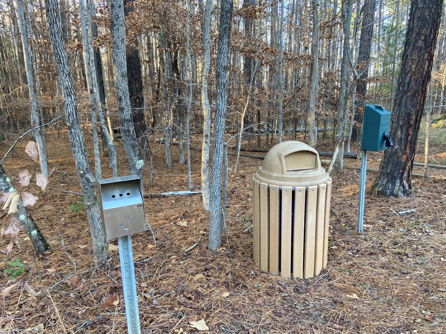 A park-like setting with a garbage can, pet waste bag dispenser, and cigarette butt disposal box