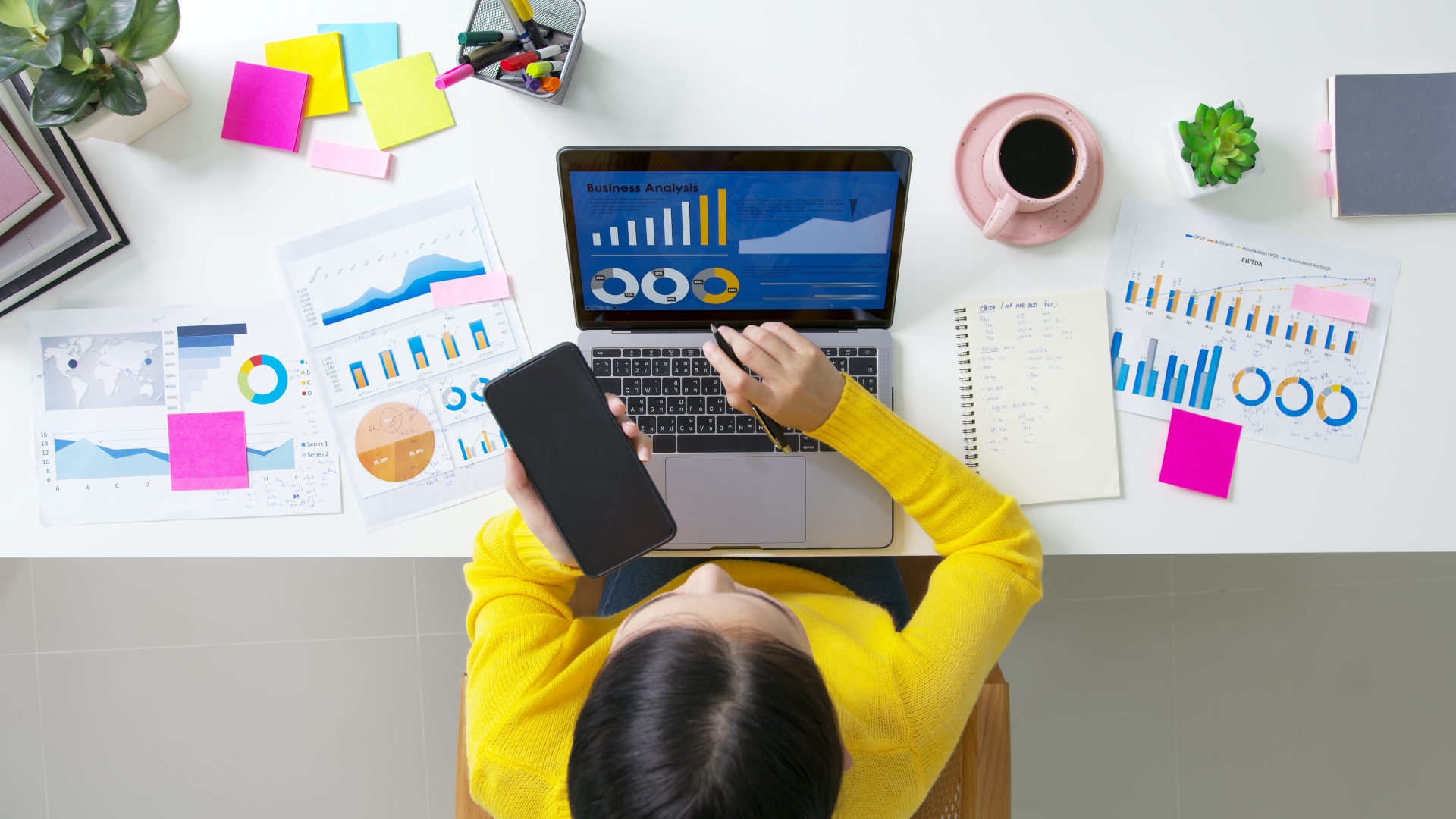 arial photo of woman looking at her computer with a screen that says "business analysis." There are pages of reports on the table around the computer. 