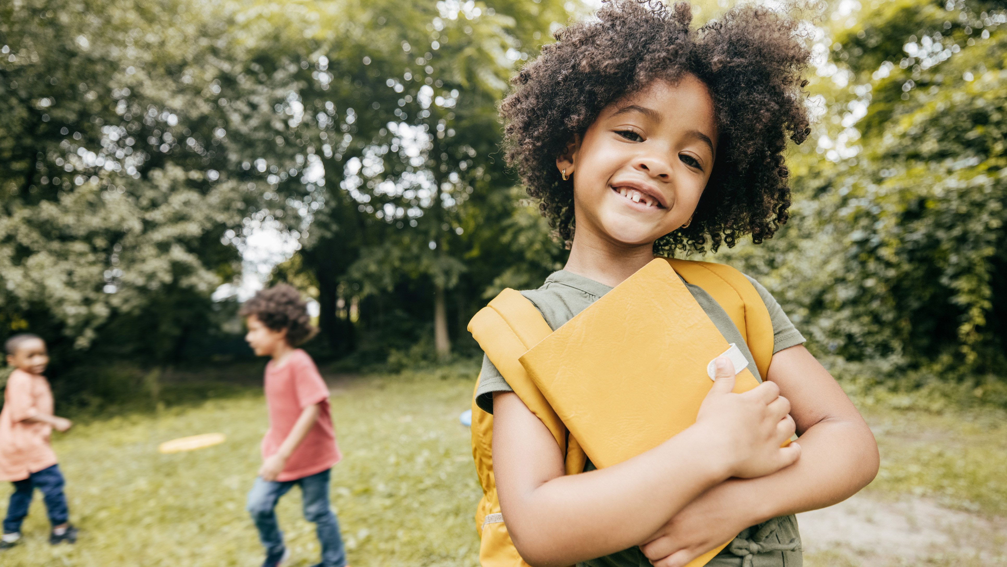 smiling child holding school books with friends playing in the background