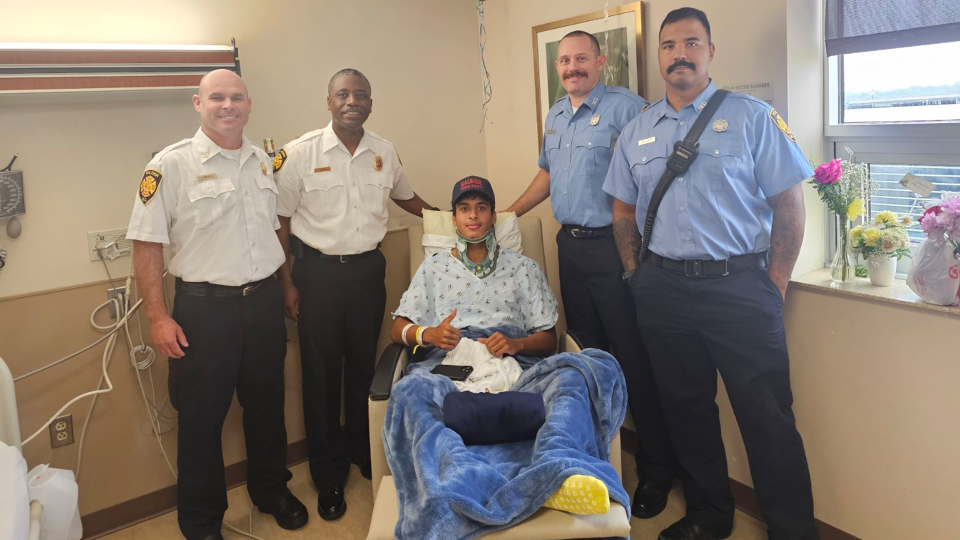 Four Raleigh firefighters in uniform gather around Bounagui who lays in hospital bed