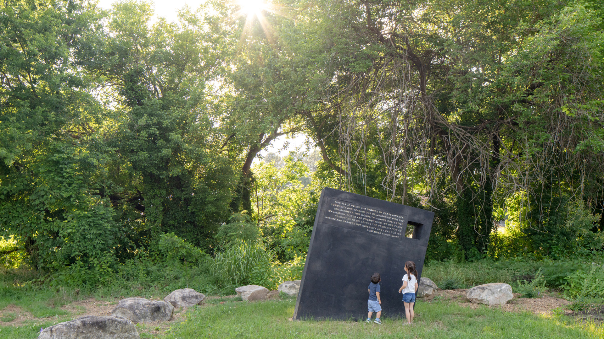 Two children standing looking at the Monolith art structure which is surrounded by grass and rocks with trees behind it. The sun is shining through the trees.