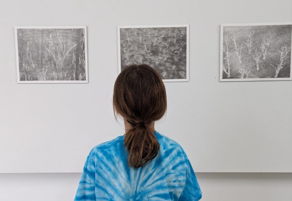 Young white girl with a long pony tail and blue tie dye shirt looking art three pencil drawings on a gallery wall. 
