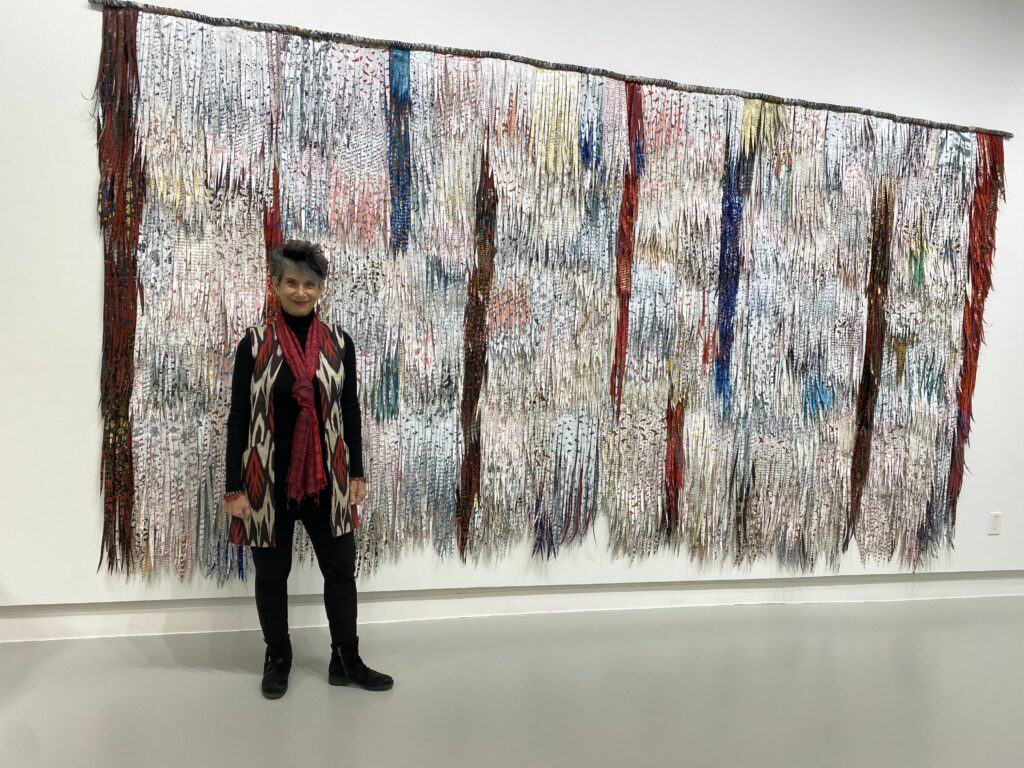 Artist, a slight, white woman with short grey hard, in front of a textured fiber wall hanging in a museum. 