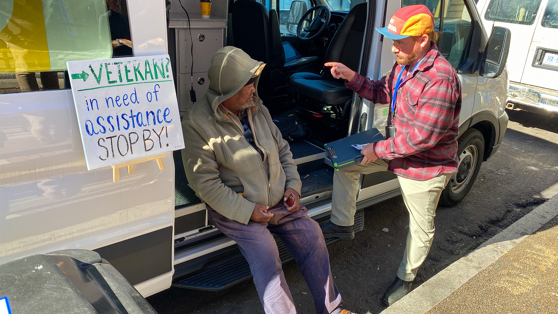 Officer Jackson talks to Army veteran Thomas Matthews as he sits on the edge of open door of van. Sign reads "Veteran? In need of assistance stop by!"