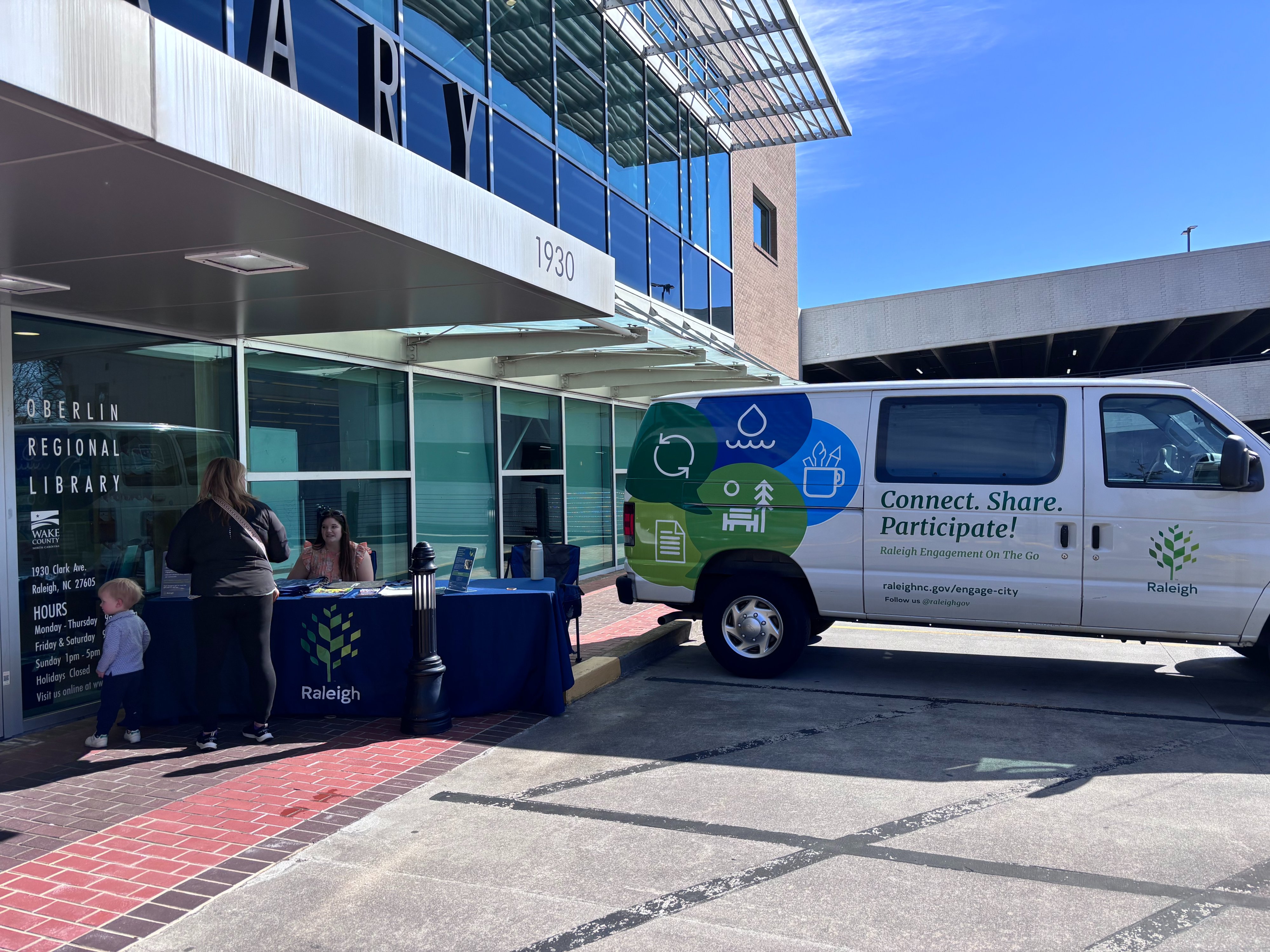 Engagement Van is parked in front of a Wake County Library location. A mother and her child speak to the City staff member at a table beside the van.