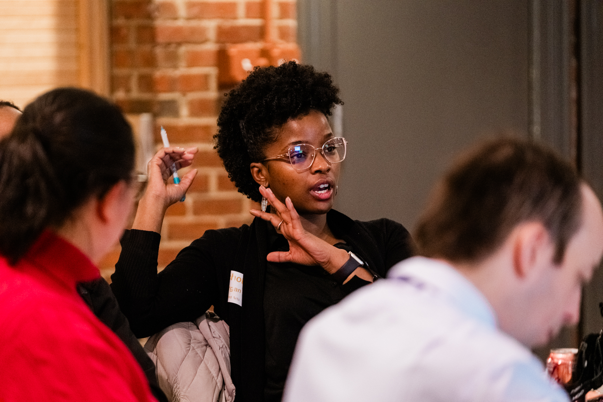 A young black women wearing a black shirt and round glasses participating in a dicussion around a table. 