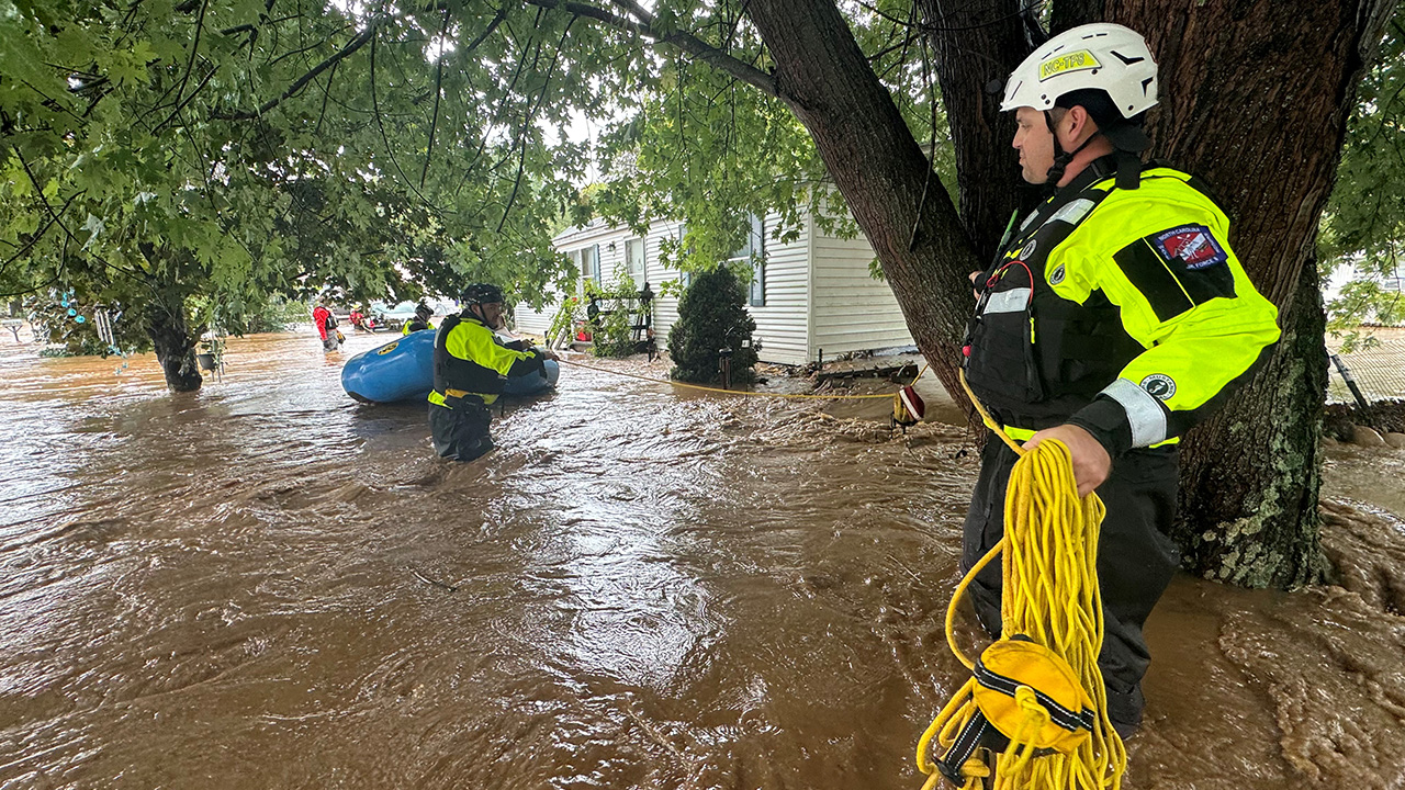 A flooded street with emergency personal standing int he water with a rope getting ready to throw it. A boat of rescue workers in the distance.