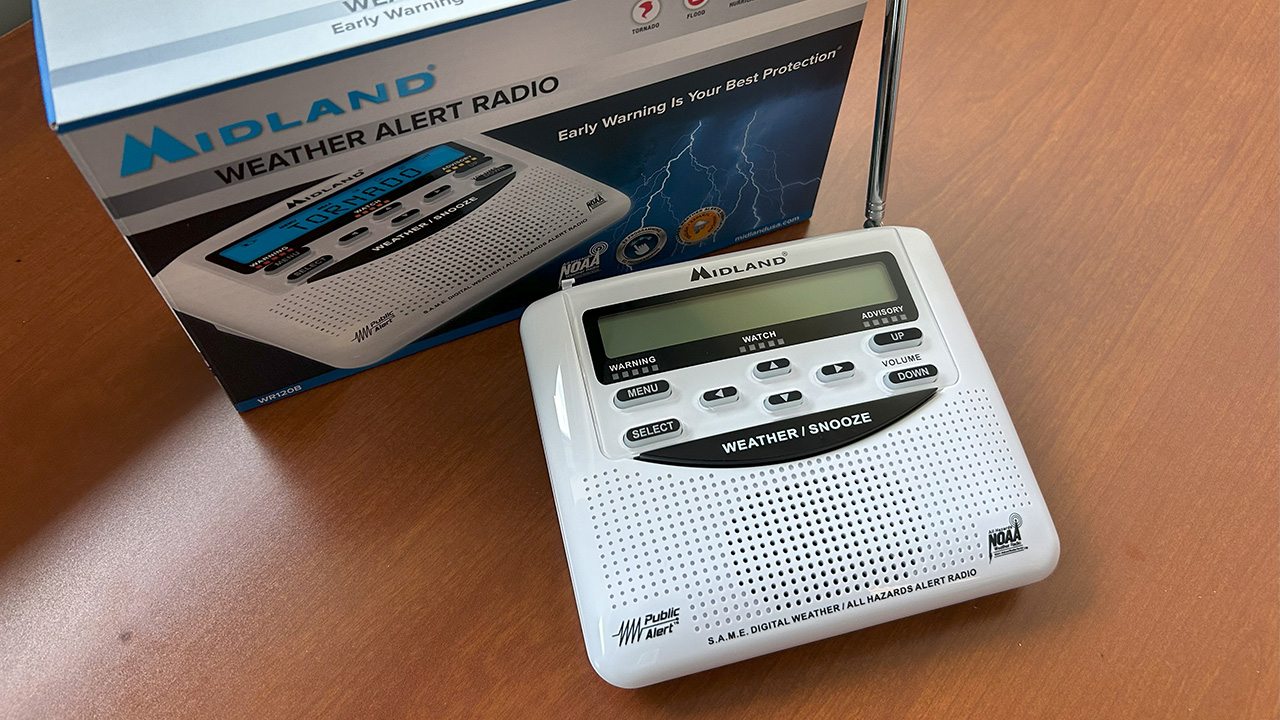 A white weather radio sitting on a wood desk with the box for the radio sitting next to the radio.