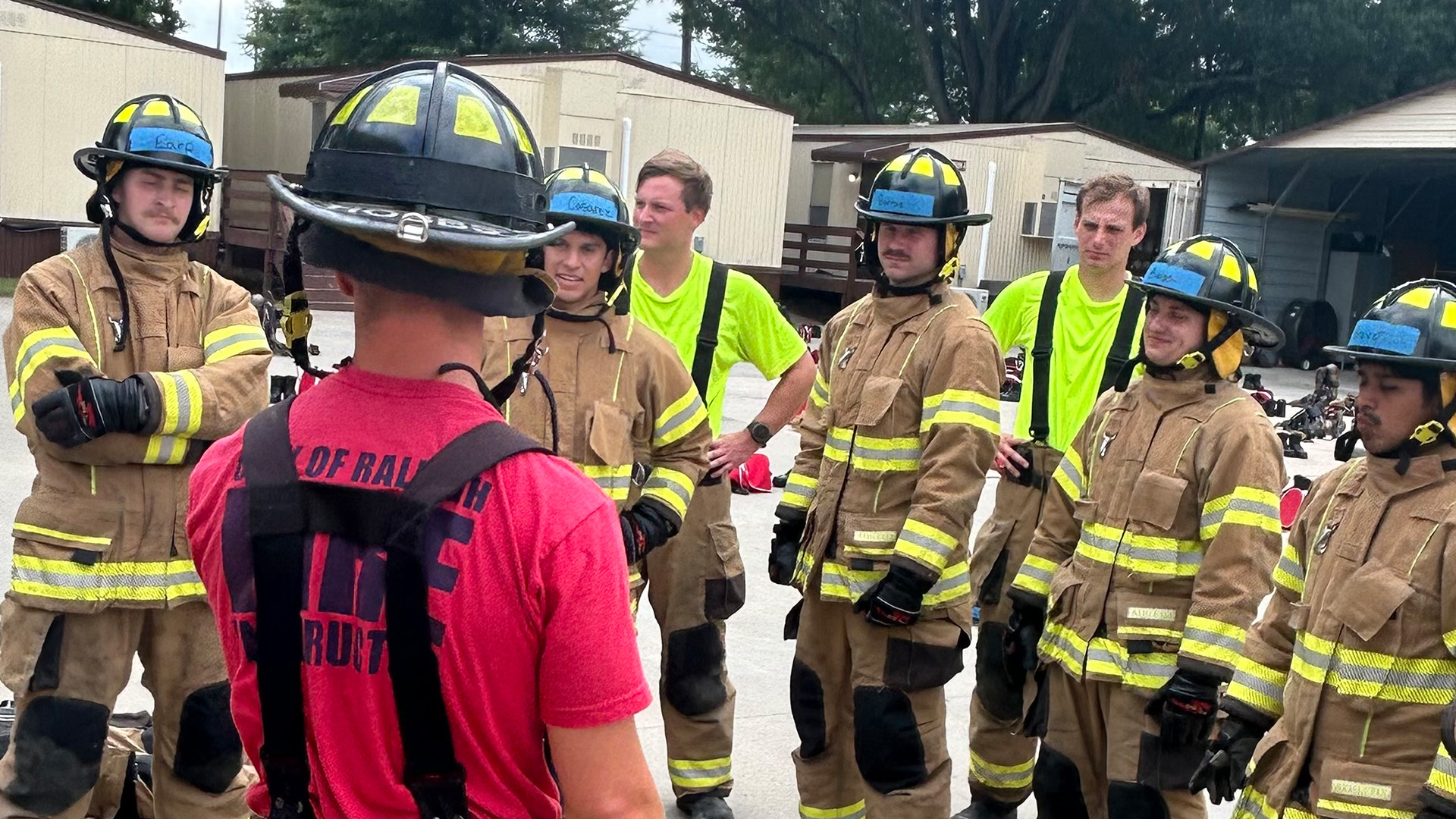 A Fire Academy insturctor in a red t-shirt talks to a group of recruits dressed in protective fire gear.