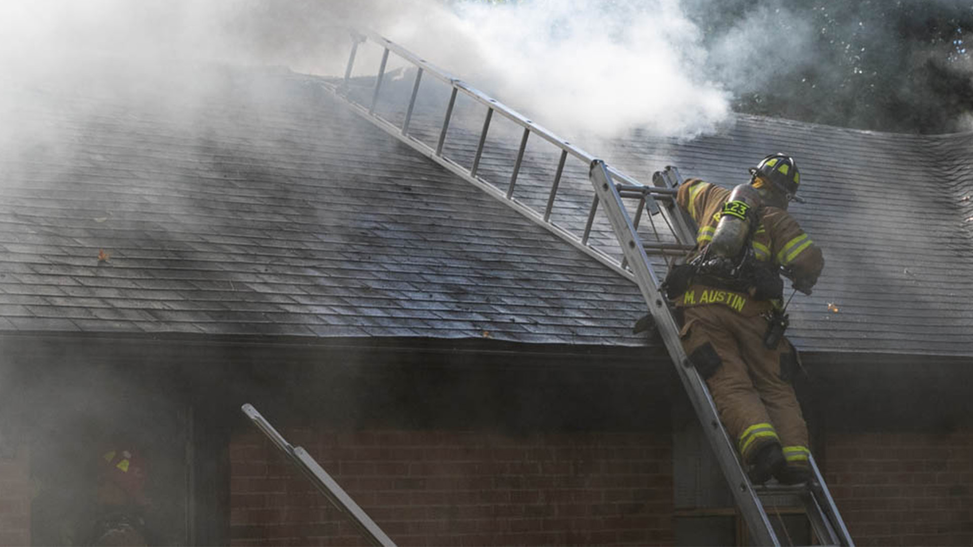 A firefighter stands on a ladder against a roof. Smoke is coming out of the roof
