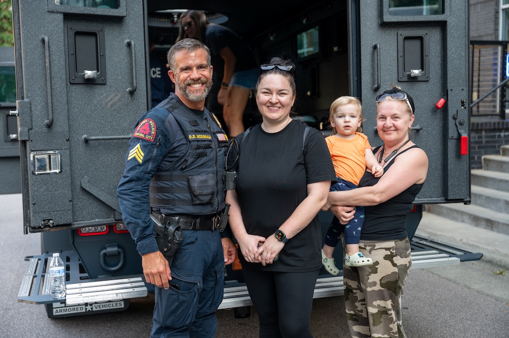 A Raleigh Police officer with two women and a small child standing in front an armored vehiclea smiling at the camera.