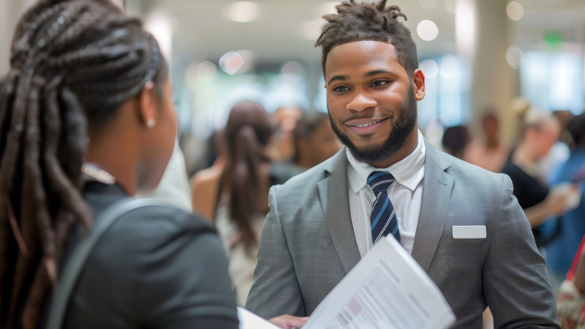 A man holding a piece of paper is talking to a woman with people in the background.