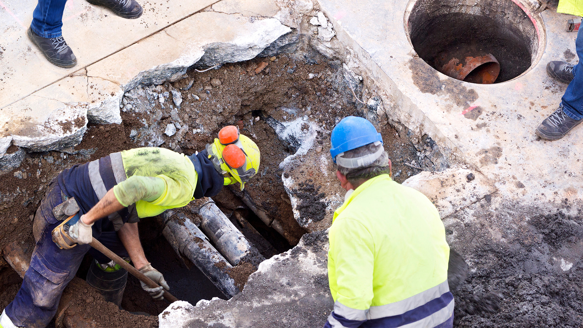 Construction works working in a hole in pavement with pipes visible.