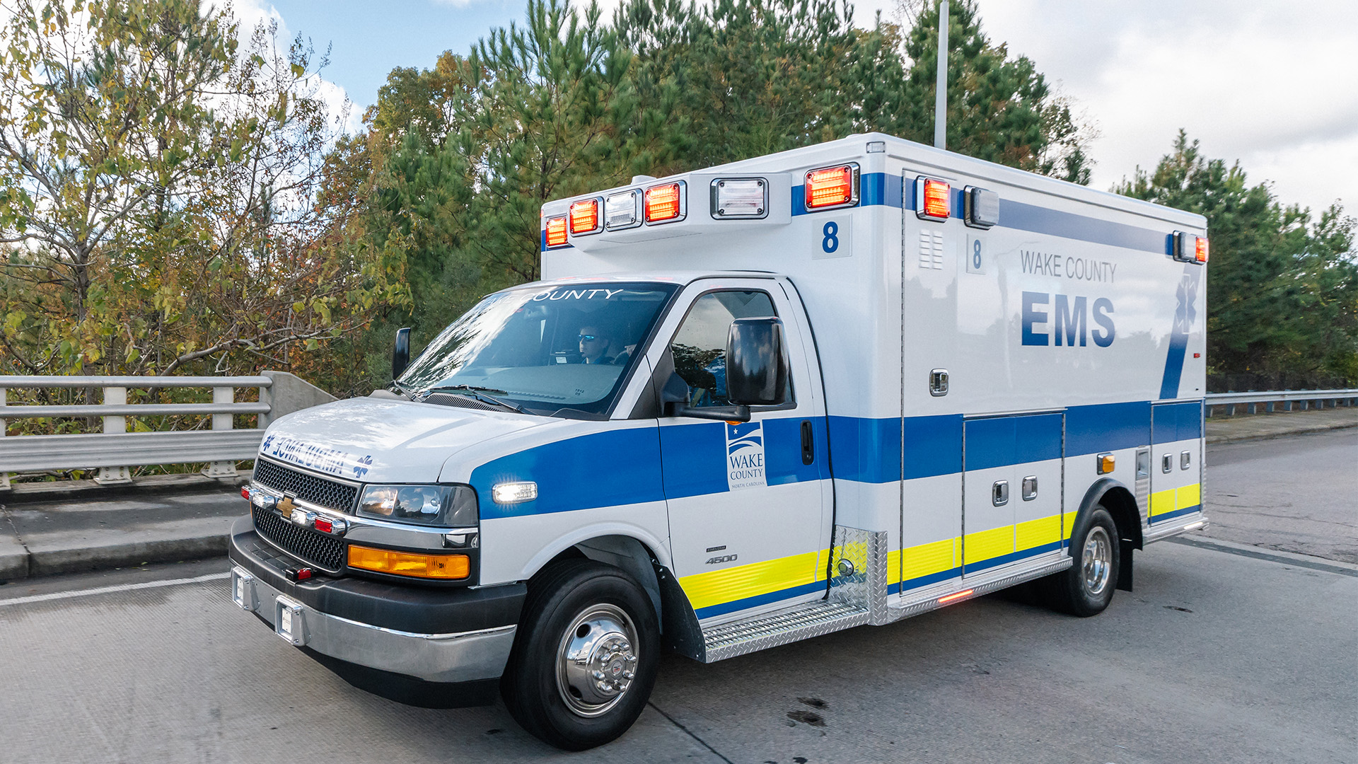 An white ambulance with a blue and yellow stripe getting ready to cross a bridge. Trees are in the background