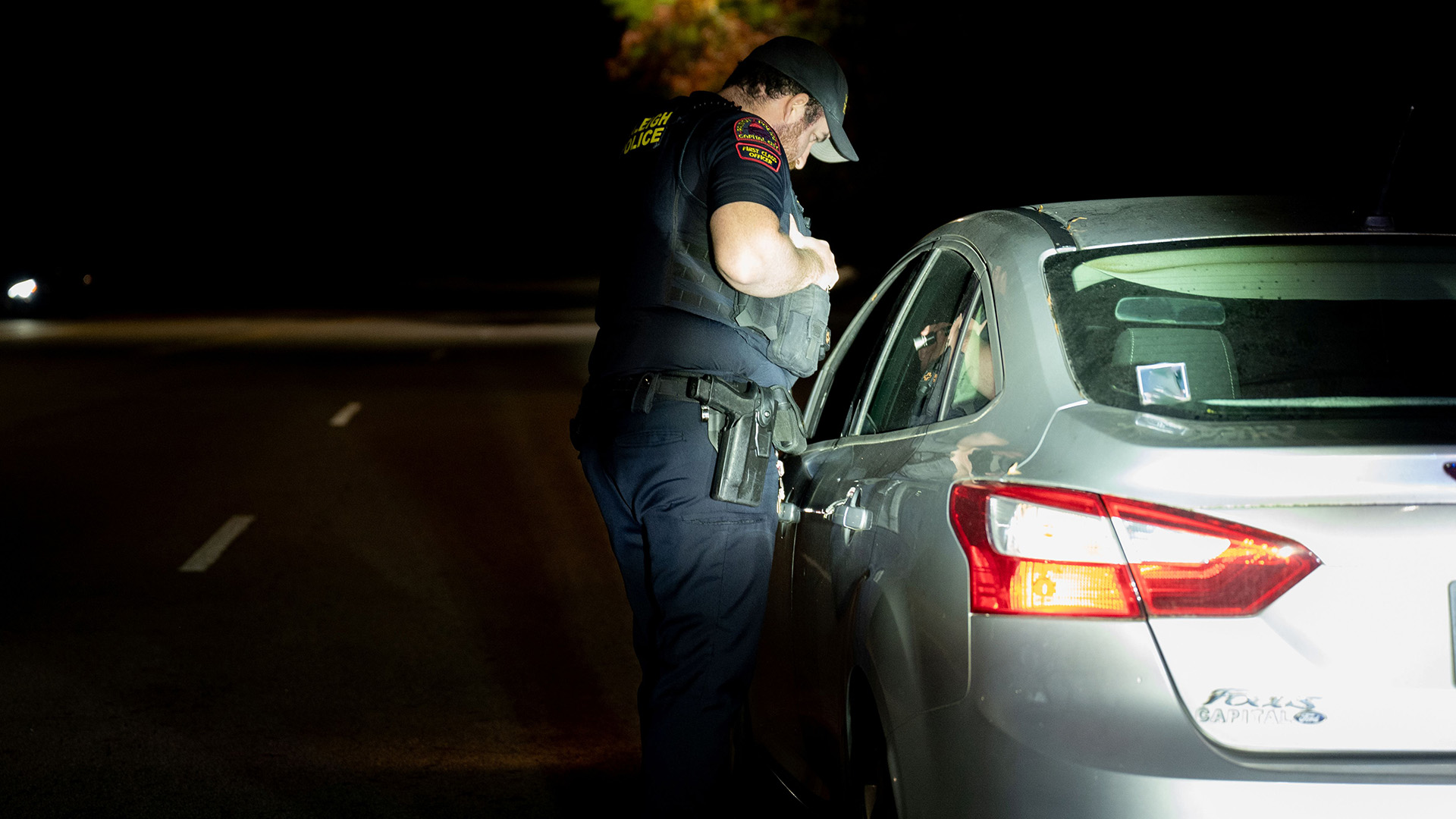At night, a Raleigh police officer stand by the driver side of a car talking with the driver.