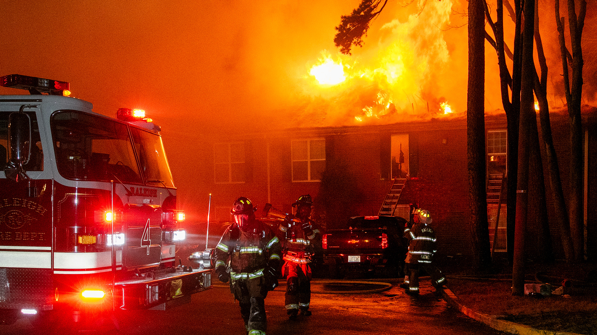 A residential building has high flames coming out of the roof at night. Raleigh firefighters are dressed in gear and working the fire. The front of the fire truck can be seen on the side of the image.