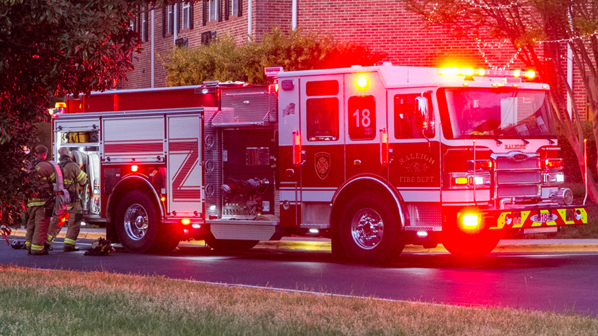 Fire truck with lights glaring and two firefighters pulling hose off the back of truck