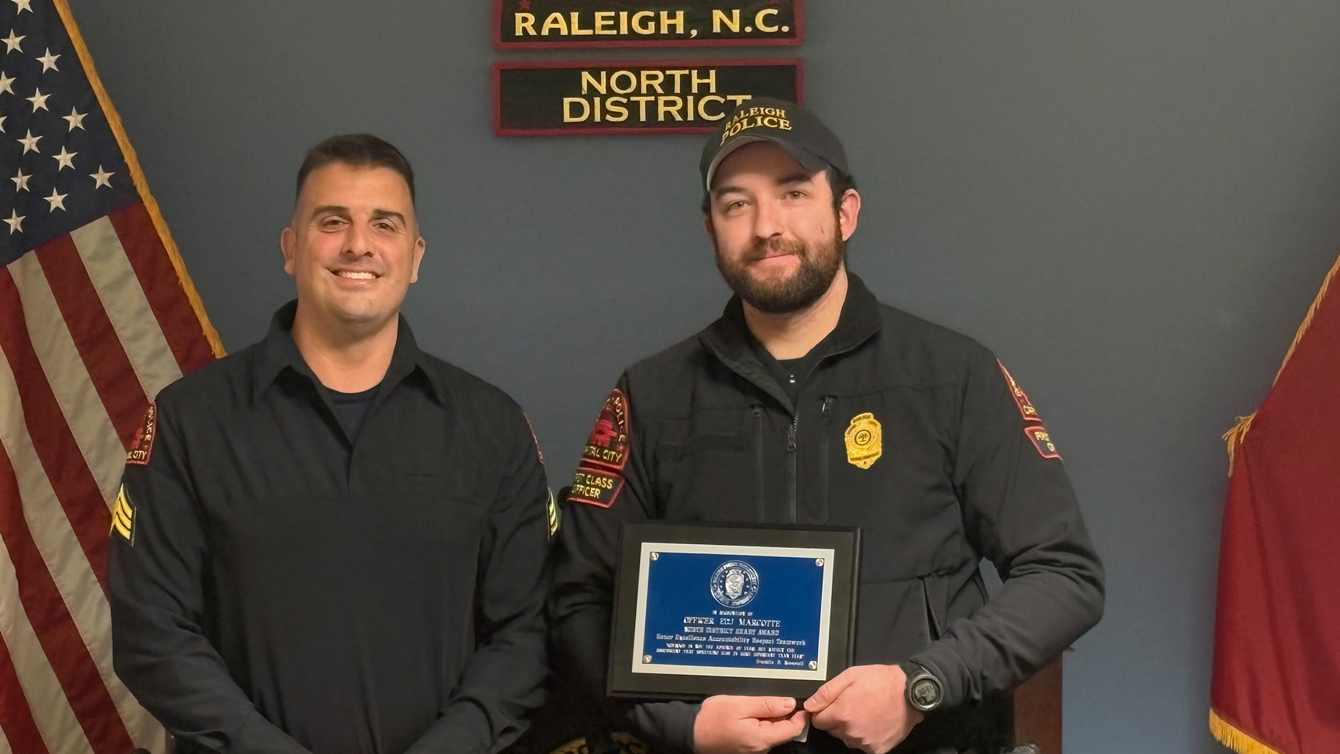 Two officers stand in front of the North District, Raleigh, NC sign. One officer holds a plaque.