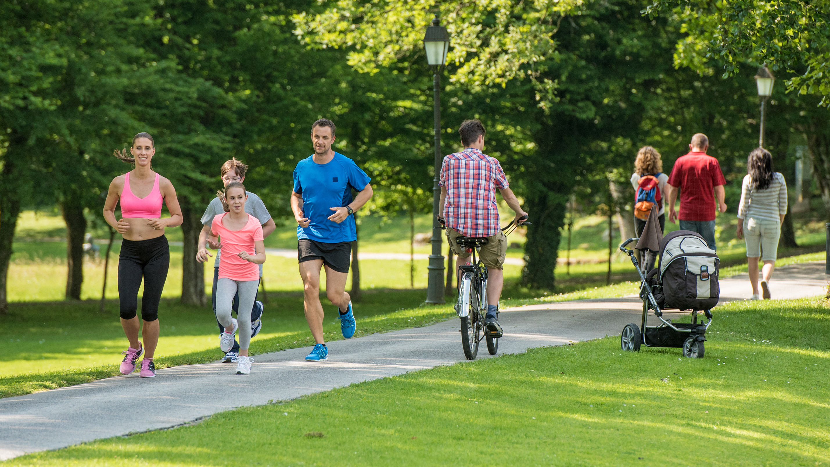 Family jogging and man cycling in park, people walking in background.