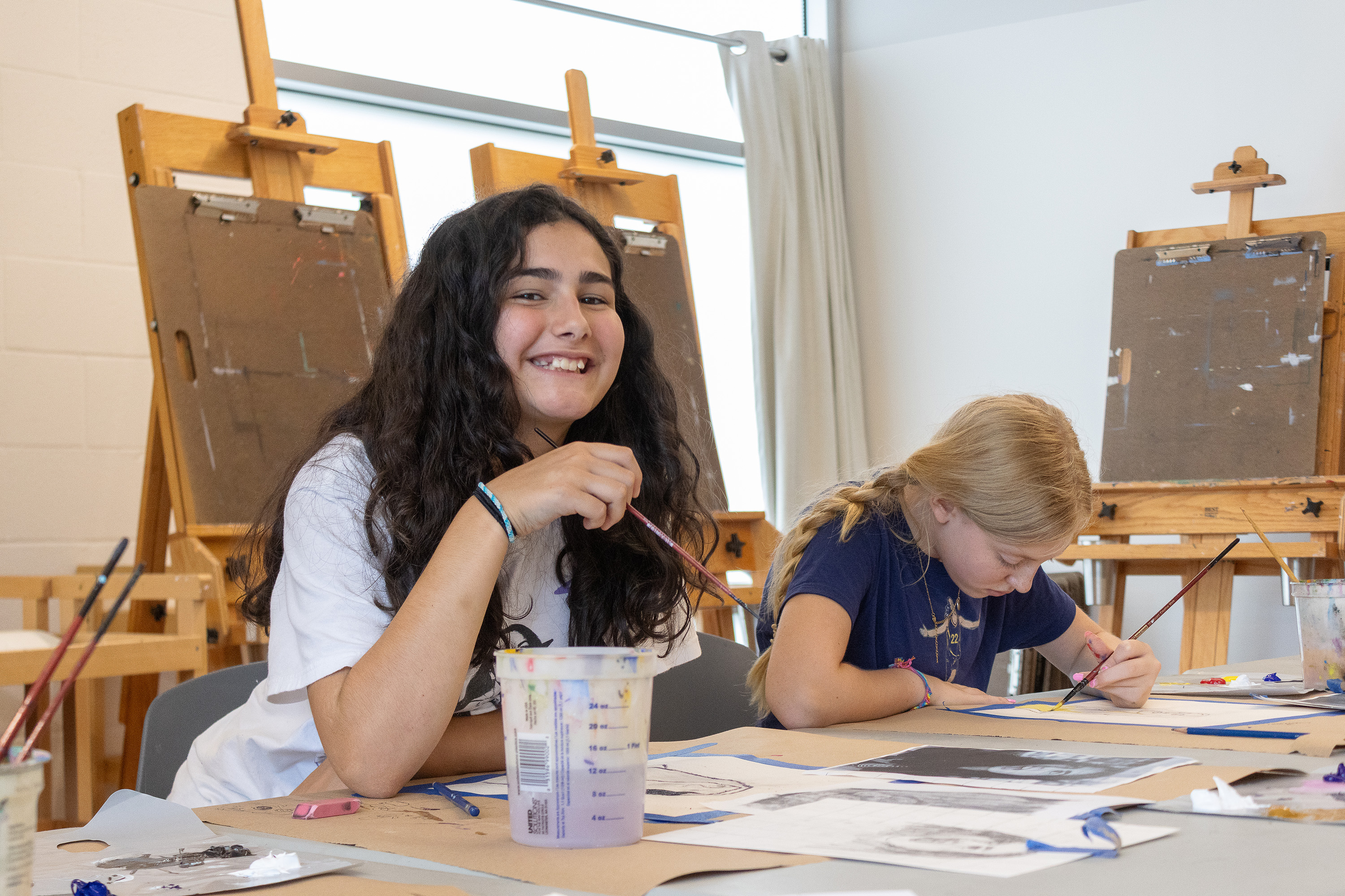 A photograph of two campers painting with acrylics at a table in a studio.