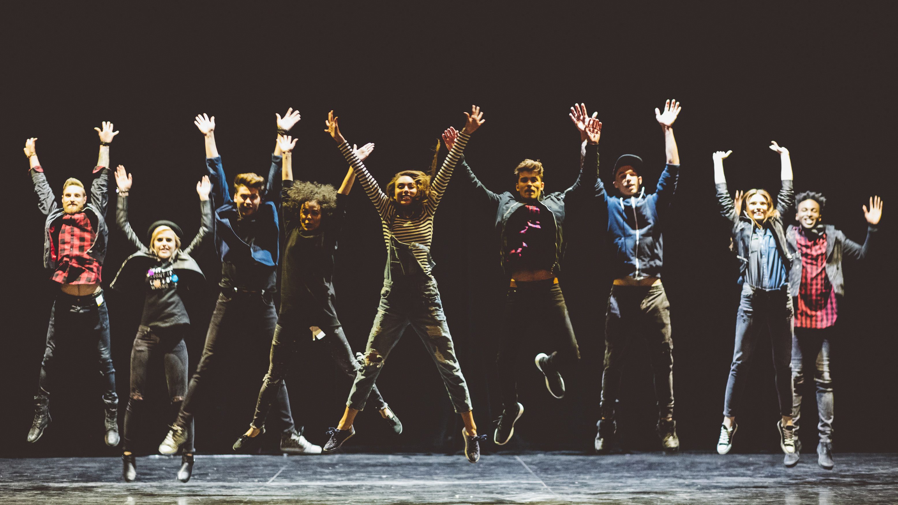 a group of young performers on stage standing in the line, jumping with raised hands