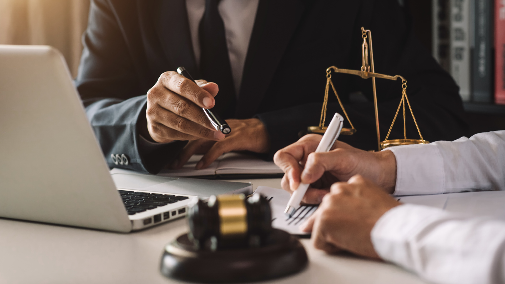 A photograph of a person signing a legal document on a desk with scales and a laptop int he background.