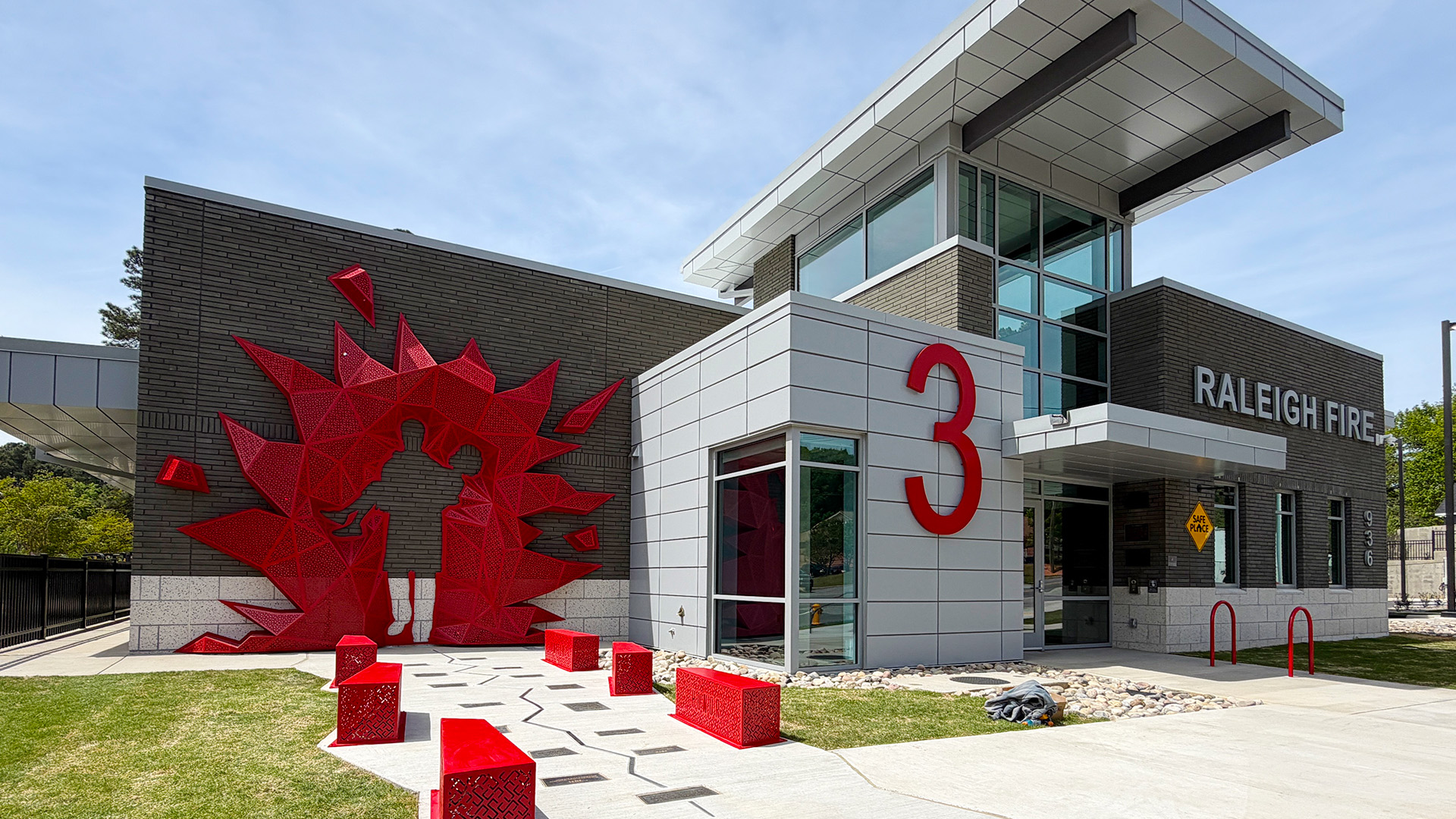 A red metal sculpture with the outline of a firefighter rescuing a woman is on the wall of a fire station building.