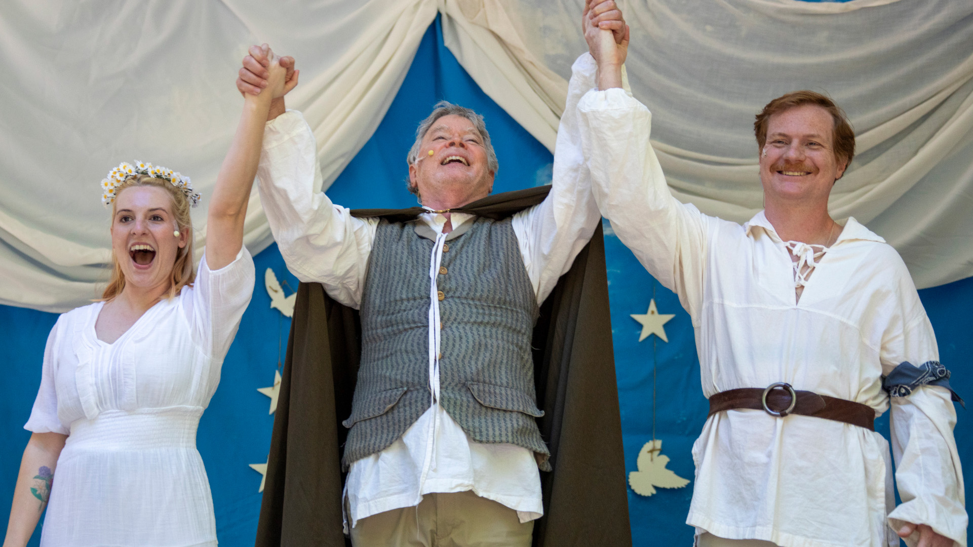 three actors on an outdoor stage joining hands and smiling