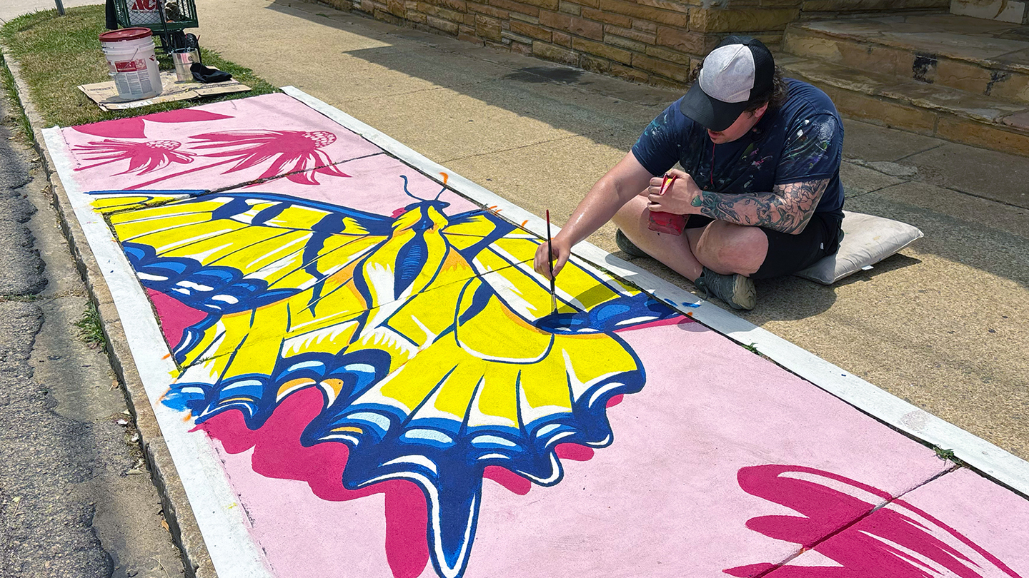 A man sits on the sidewalk painting a large monarch butterfly mural in yellows and pinks.