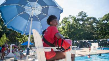 female lifeguard sitting in lifeguard chair at biltmore pool smiling with view of pool in the background