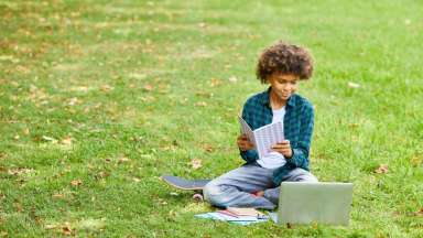 Teenage boy sitting out in grass holding notebook and looking at laptop