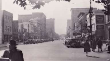 Historic photo of downtown street with shops lining street and people walking around