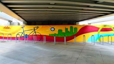 A mural by artist JP Jermaine Powell on wall behind metal bike racks at Raleigh Union Station. The mural features a bicycle, a sun painted around a light fixture, and a silhouette of the city skyline with colorful wavy lines.
