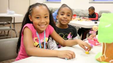 Two girls sitting at table holding doll and smiling