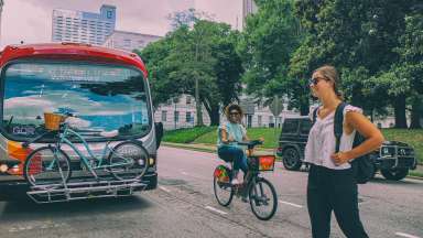 A GoRaleigh bus with a blue bike on the front rack. Cityscape and trees in the background. A woman smiles while riding a Citrix Cycle beside the bus as another woman crosses in front with a backpack