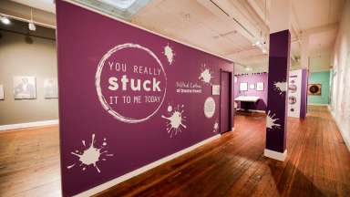 View of purple exhibition display board showing title of "You really stuck it to me today: The Political Cartoons of Dwane Powell"