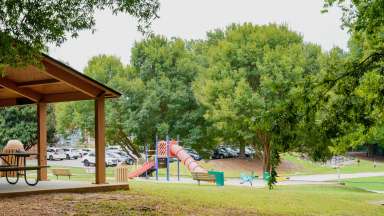 Wide shot of park with picnic shelter on left, playground and sand diggers in distance. under canopy of trees