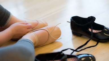 Child's hand and feet in ballet shoes with tap shoes shown nearby