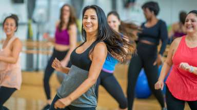 A multi-ethnic group of adult women are dancing in a fitness studio. They are wearing athletic clothes. An Ethnic woman is smiling while dancing in the foreground.