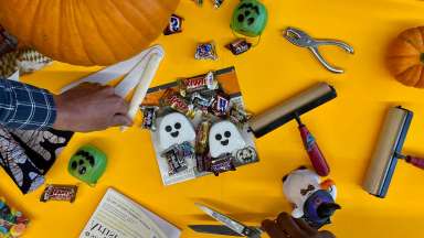 An assortment of candy, Halloween decor, print making tools are scattered on a yellow backdrop. One hand is seen picking up a tool while another hand holds scissors.