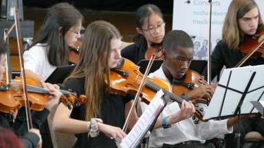 A group of young musicians are seated reading music and playing violins during a performance