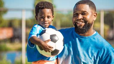 A smiling man holds his young son, who is holding a soccer ball.