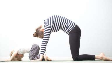 A mother and a child kneeling and bending forward with a rounded back, practicing yoga together.