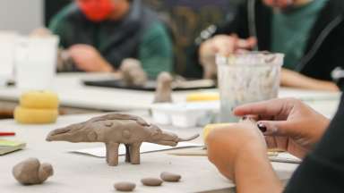 A teen sits at a table with others and constructs a dinosaur out of beige colored clay