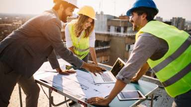 Men and women stand around table with blue prints at construction site