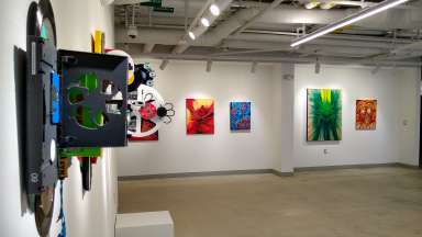 The white walls of the gallery exhibition space at Pullen Arts Center including colorful hanging artworks by Leatha Koefler and Michael Bennett